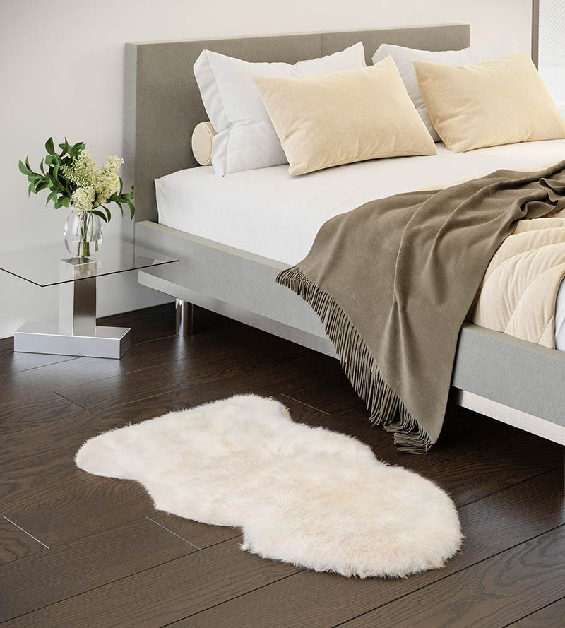 Ivory sheepskin rug on a wooden floor in front of a bed with pillows and a blanket.