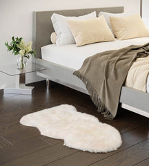 Ivory sheepskin rug on a wooden floor in front of a bed with pillows and a blanket.